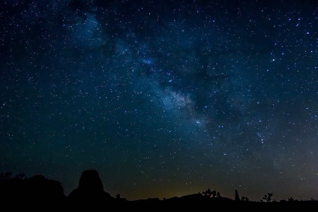 The Milky Way arching over a dark landscape with a faint silhouette of trees on the horizon.