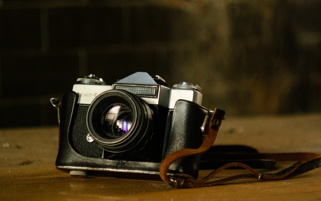 A hand holding a vintage 35mm film camera, with several rolls of film and a contact sheet resting on a wooden table.