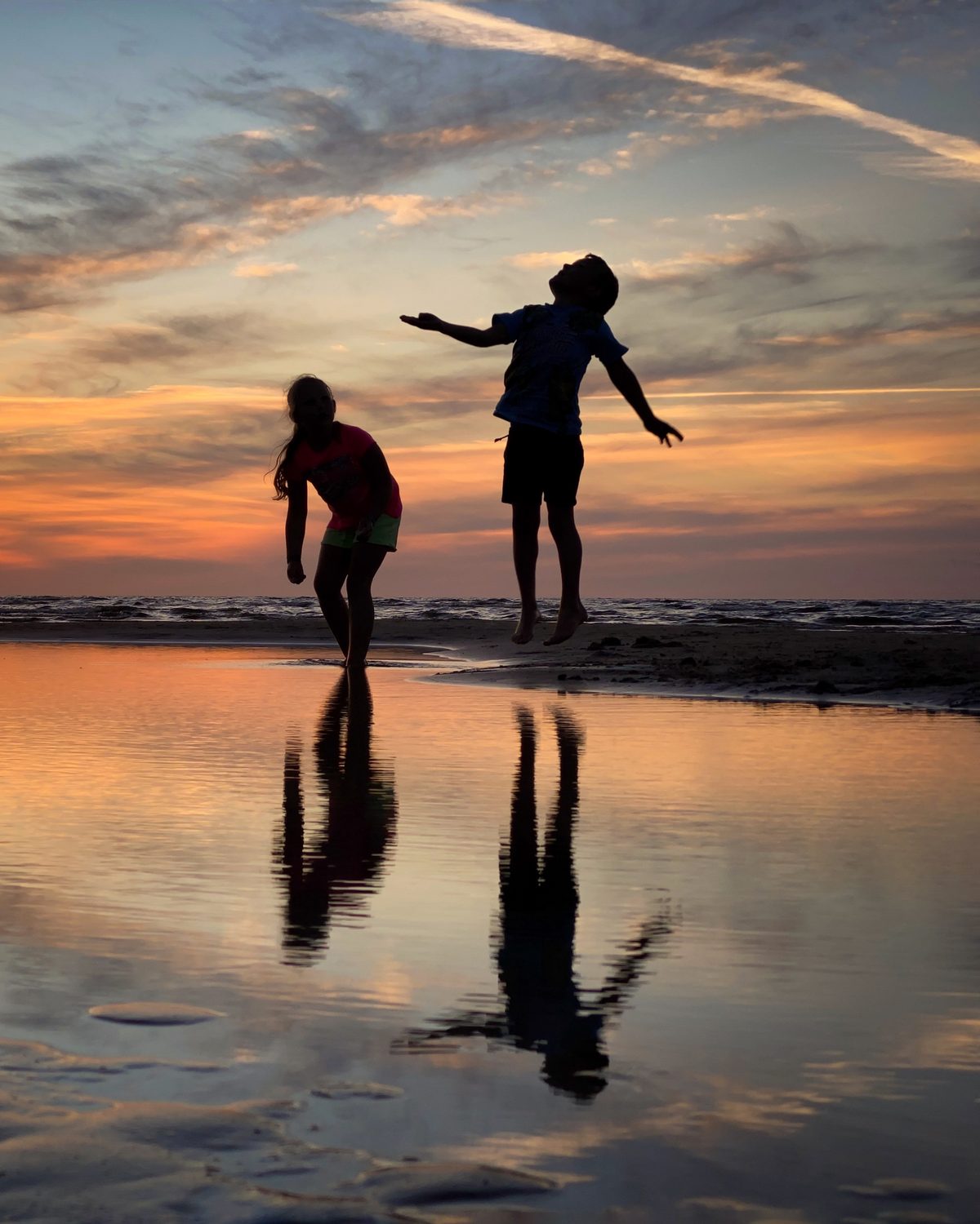 Two children playing on the beach at sunset, reflected in the wet sand.
