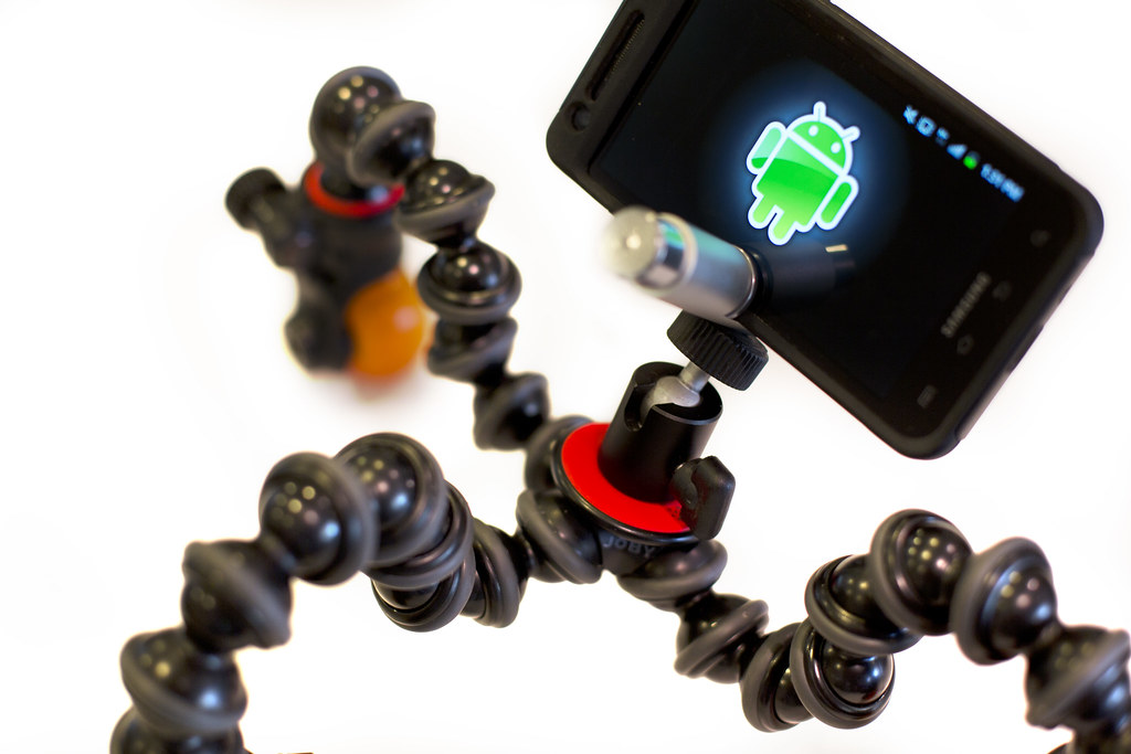 A smartphone mounted on a small tabletop tripod with a clip-on lens and a tiny LED light beside it.