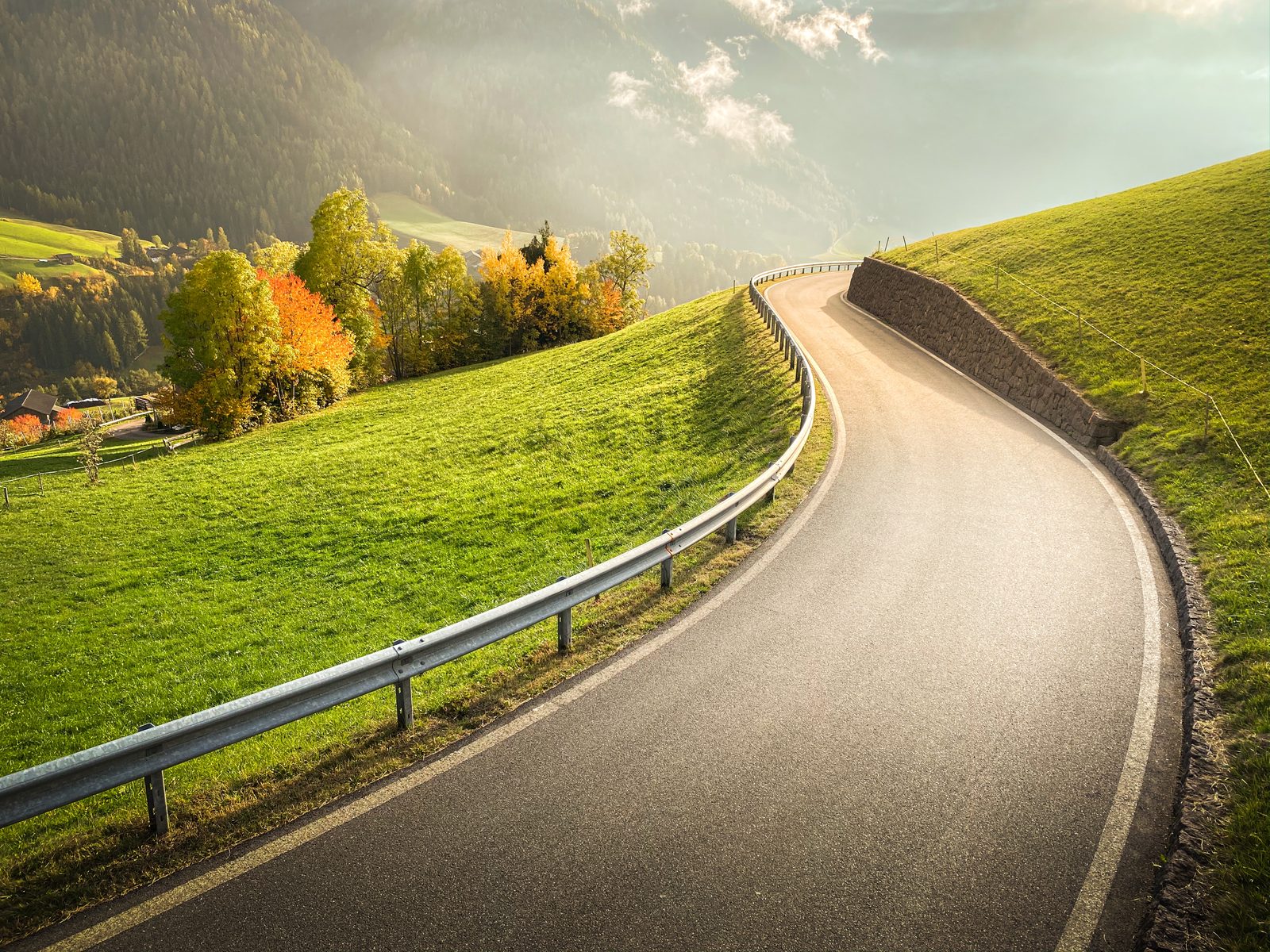 A narrow mountain road curving through green alpine slopes and autumn trees.