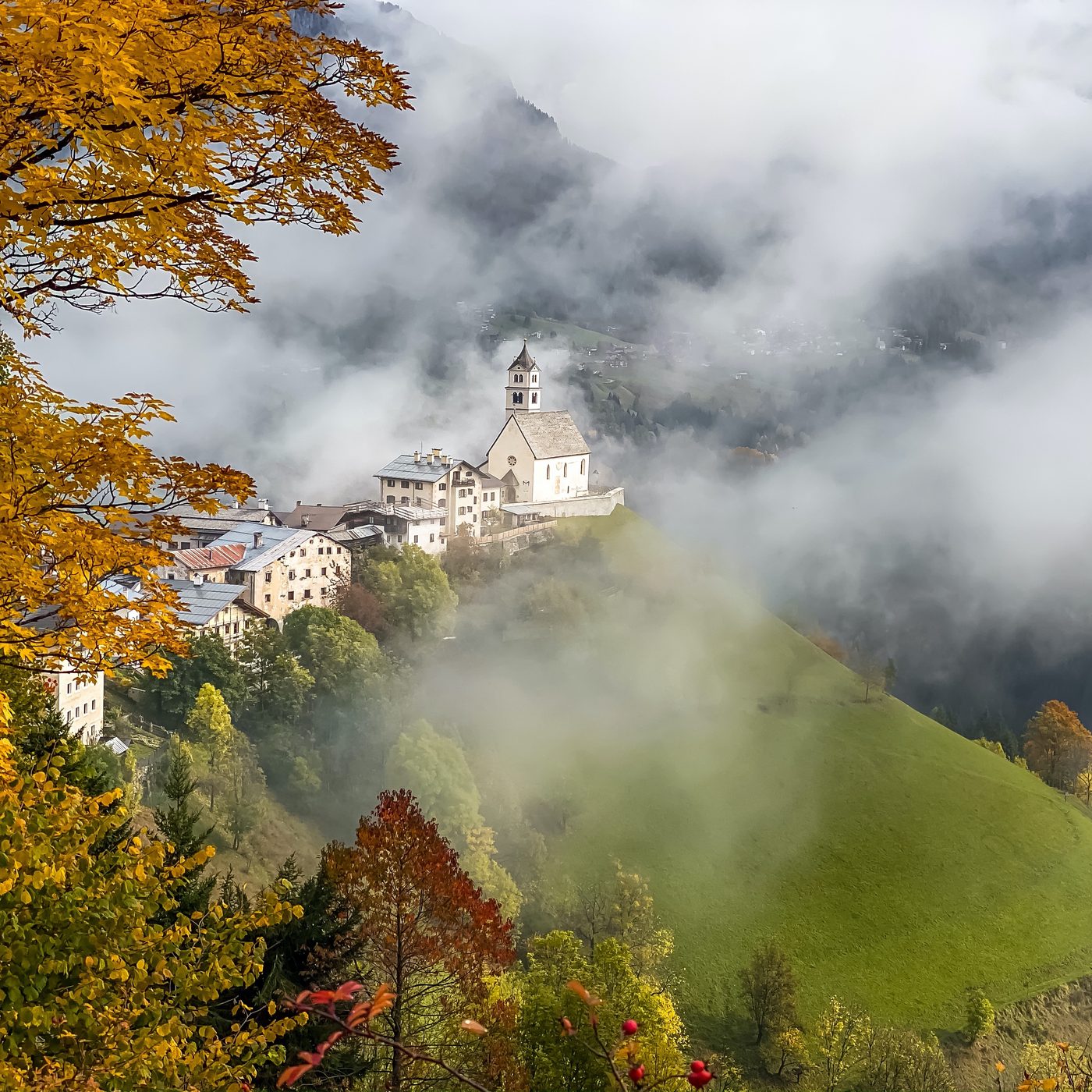 A hilltop village and church emerging from fog, framed by golden autumn leaves.